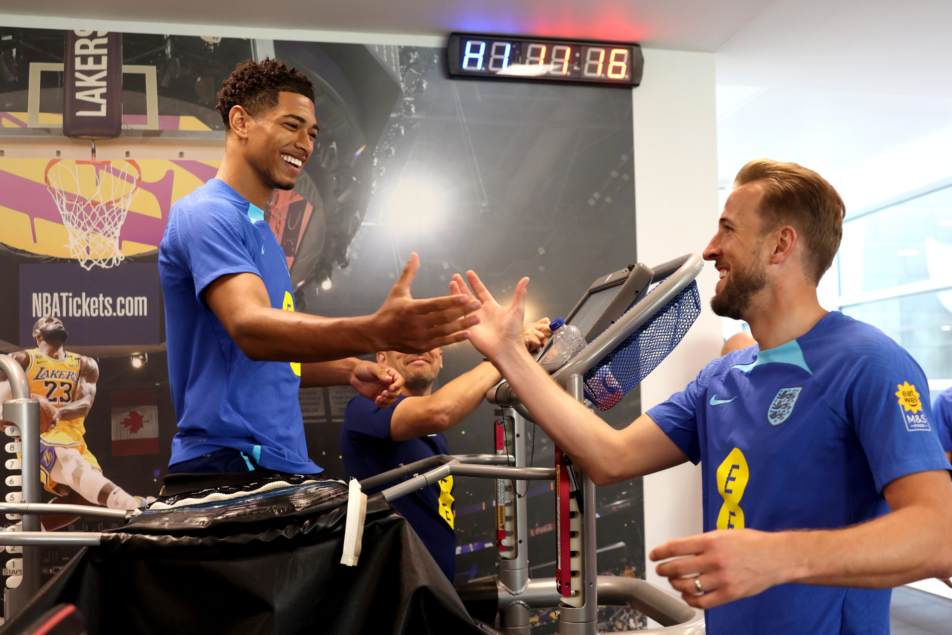  Harry Kane and Jude Bellingham, both wearing blue England training shirts, shake hands while walking on treadmills in the gym.